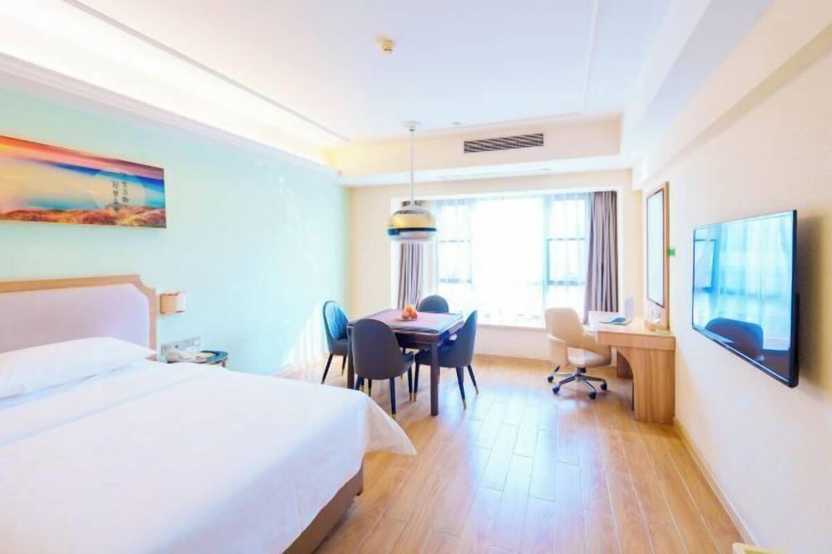 A hotel room with a large bed, a desk with a chair, a dining table with chairs, a TV mounted on the wall, and a painting on the wall.