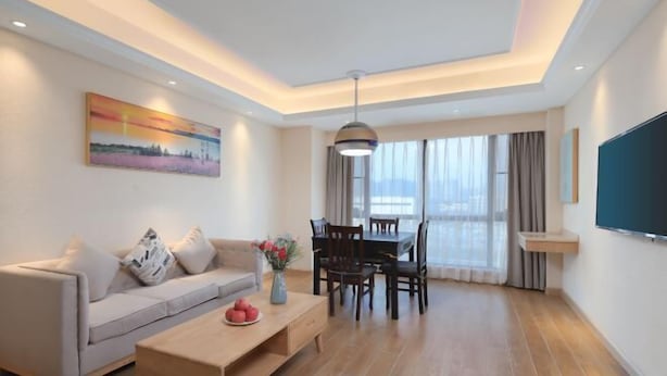 A modern living room with a sofa, coffee table, dining table, and chairs. There is a painting on the wall, a TV mounted on the wall, and a large window with curtains. - Vienna 3 Best Hotel (Changsha Wangcheng Economic Development Zone) (Changsha)
