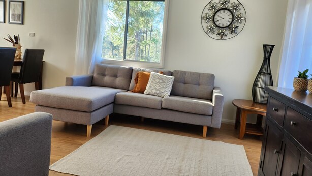 Un salon moderne avec un canapé d’angle gris, un tapis blanc, un coin repas comprenant une table et des chaises, une horloge murale décorative et une fenêtre donnant sur des arbres.