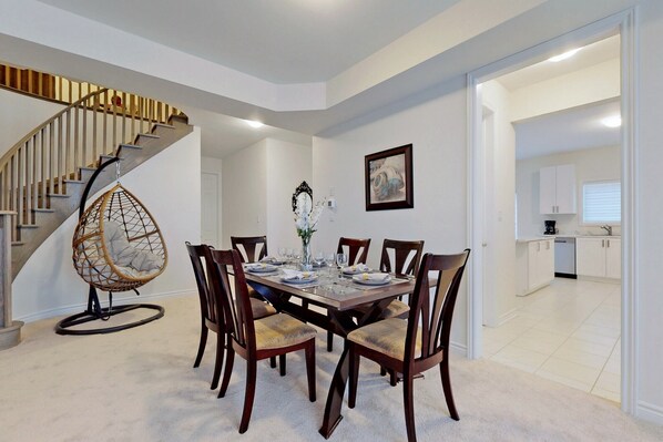 A dining area with a wooden table set for six, surrounded by wooden chairs. A unique hanging chair is positioned to the left of the table. A staircase with wooden steps and a metal railing is visible in the background.