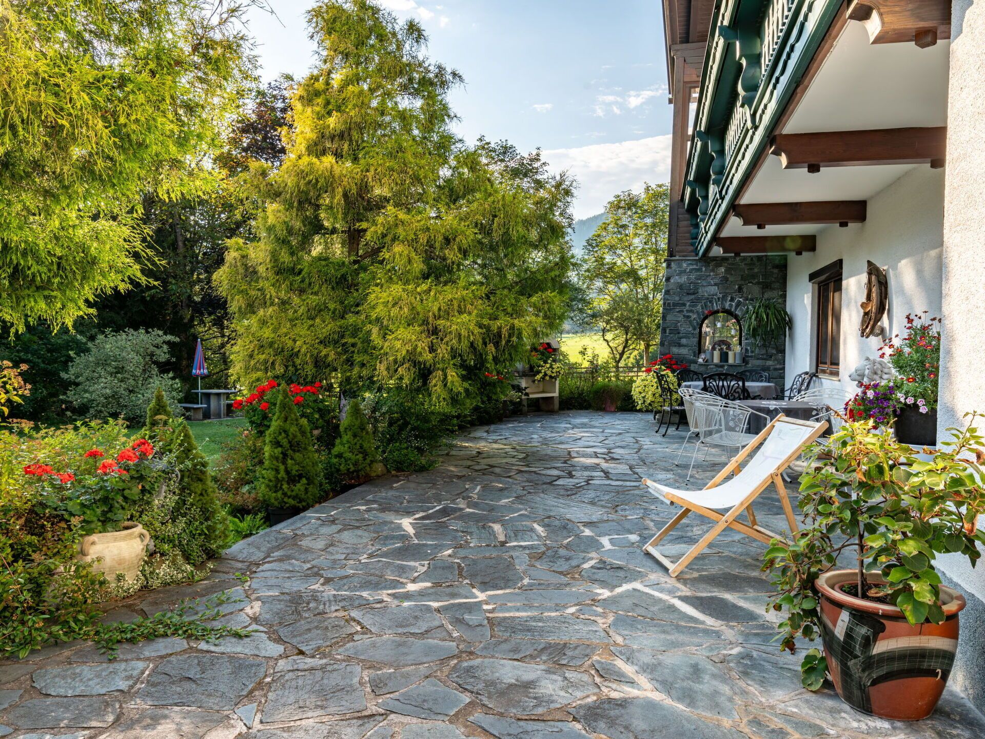 Une terrasse dallée de pierres, dotée de sièges extérieurs, entourée d’une végétation luxuriante et offrant une vue sur une montagne.