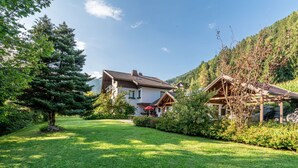 Une maison de style chalet, dotée d’un toit en pente, entourée d’une végétation luxuriante et avec une chaîne de montagnes en toile de fond.