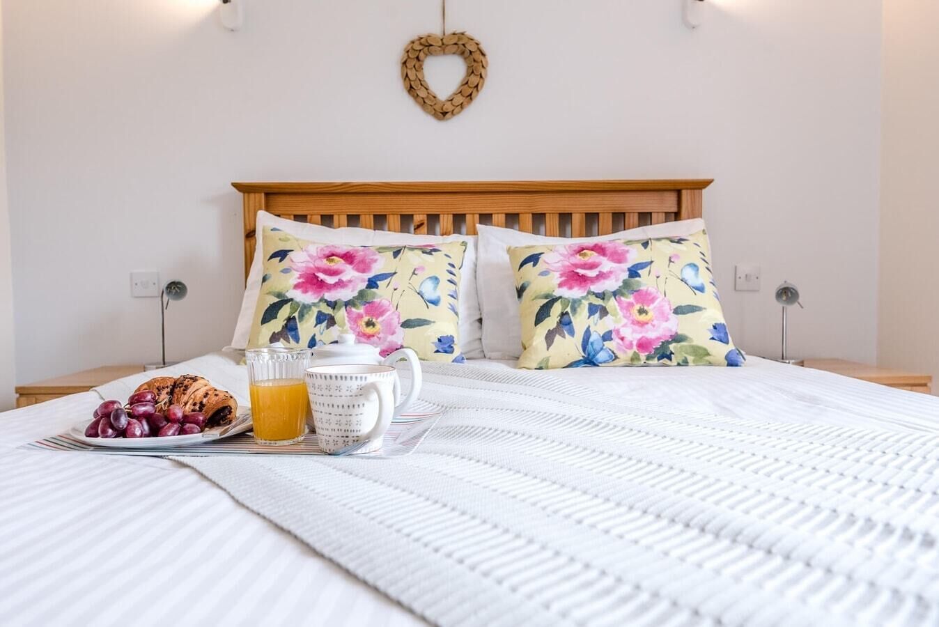 Uma cama bem arrumada com almofadas florais, um prato de uvas, um croissant e um copo de suco de laranja.