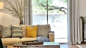 A living room with a sofa, a coffee table, and a chair. There is a window with curtains, a vase with branches, and a brick wall outside. - The Courtyard on Pio Decimo  (Tucson)