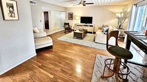 A spacious living room with hardwood flooring, a ceiling fan, a television, and a dining area with a bar stool. - The Courtyard on Pio Decimo  (Tucson)
