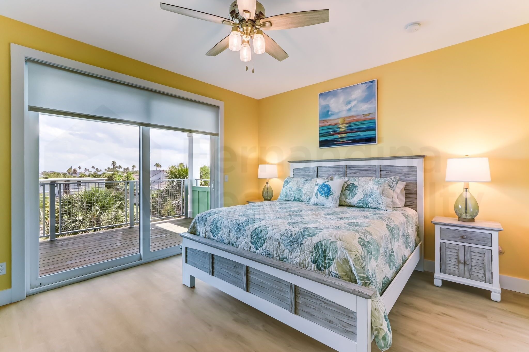 A bedroom with a large bed, two bedside tables, a ceiling fan, and a view of the outdoors.