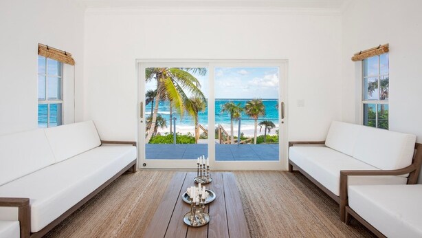 Una habitación con vista a la playa, sofás blancos, una mesa de centro de madera y una puerta con paneles de vidrio.