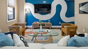Un salón moderno con un televisor grande montado en una pared azul, dos sillones, una mesa de centro de vidrio y un sofá con cojines estampados.