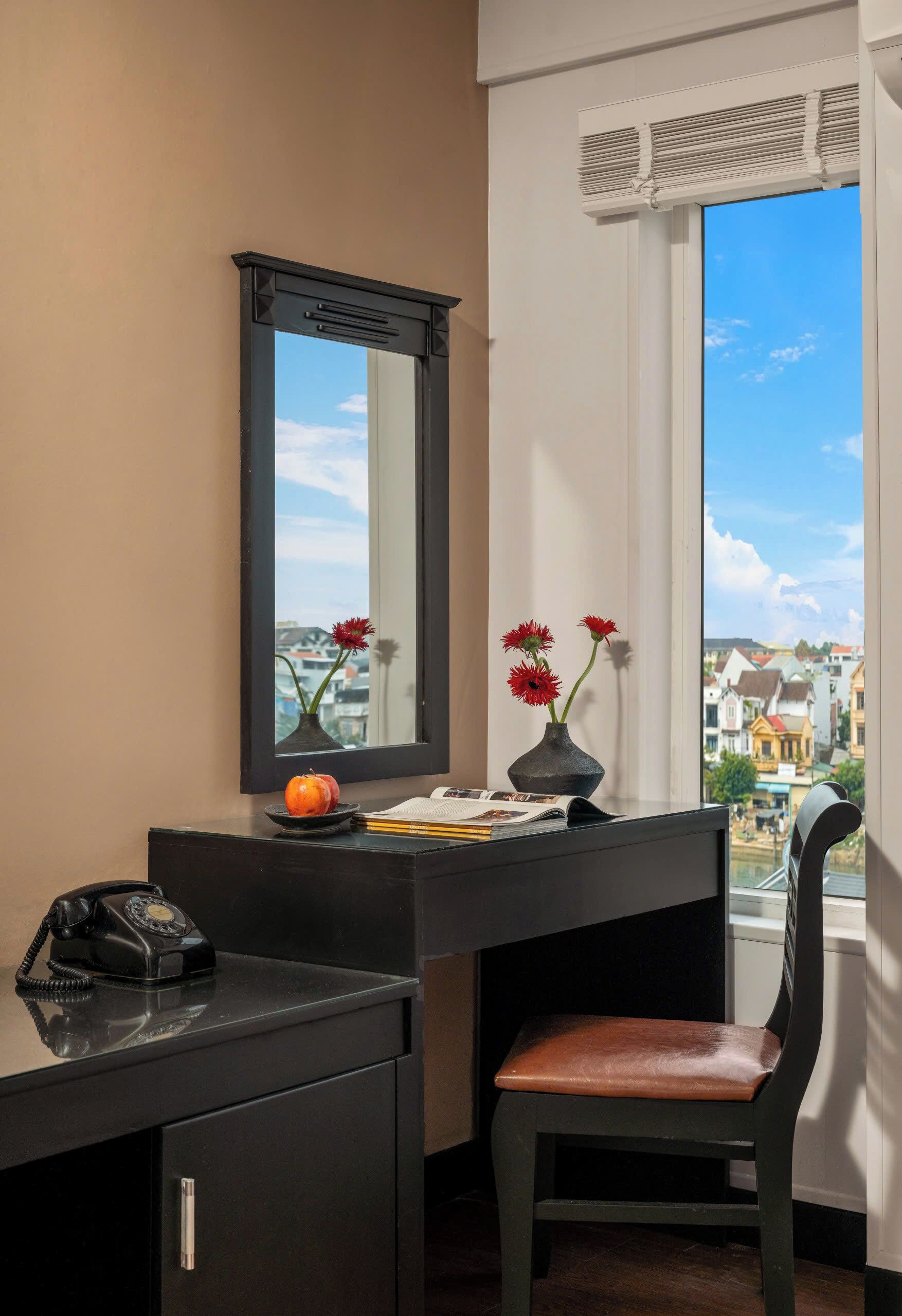 Une pièce comprenant un bureau, une chaise, un téléphone, un vase avec des fleurs et une vue sur des immeubles par la fenêtre.