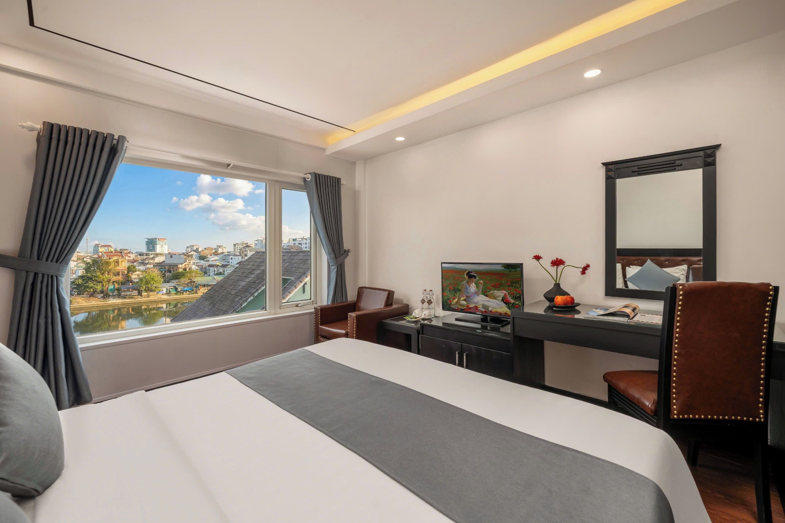 A hotel room with a bed, a desk, a chair, a television, and a view of a cityscape.