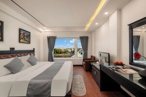A hotel room with a bed, a desk, a chair, a window with a view of the city, and a painting on the wall.