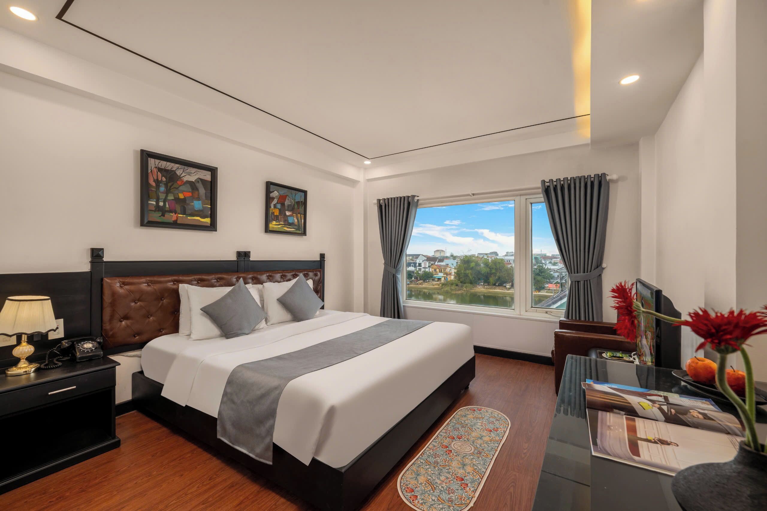 Habitación de hotel con cama, mesita de noche, lámpara, ventana con cortinas y una obra de arte en la pared.
