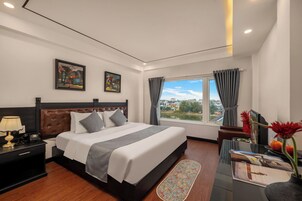 Habitación de hotel con cama, mesita de noche, lámpara, ventana con cortinas y una obra de arte en la pared.