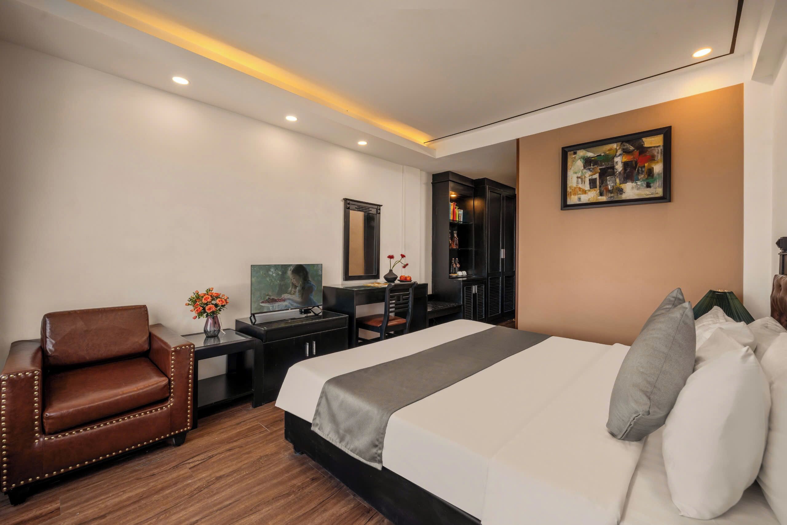 A hotel room with a large bed, a desk with a computer, a chair, a bookshelf, and a painting on the wall.