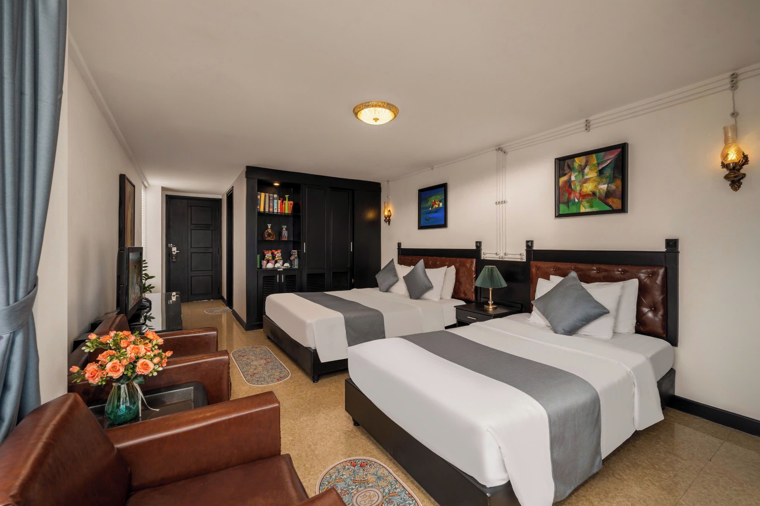 A hotel room with two beds, a desk, a chair, a bookshelf, and artwork on the walls.