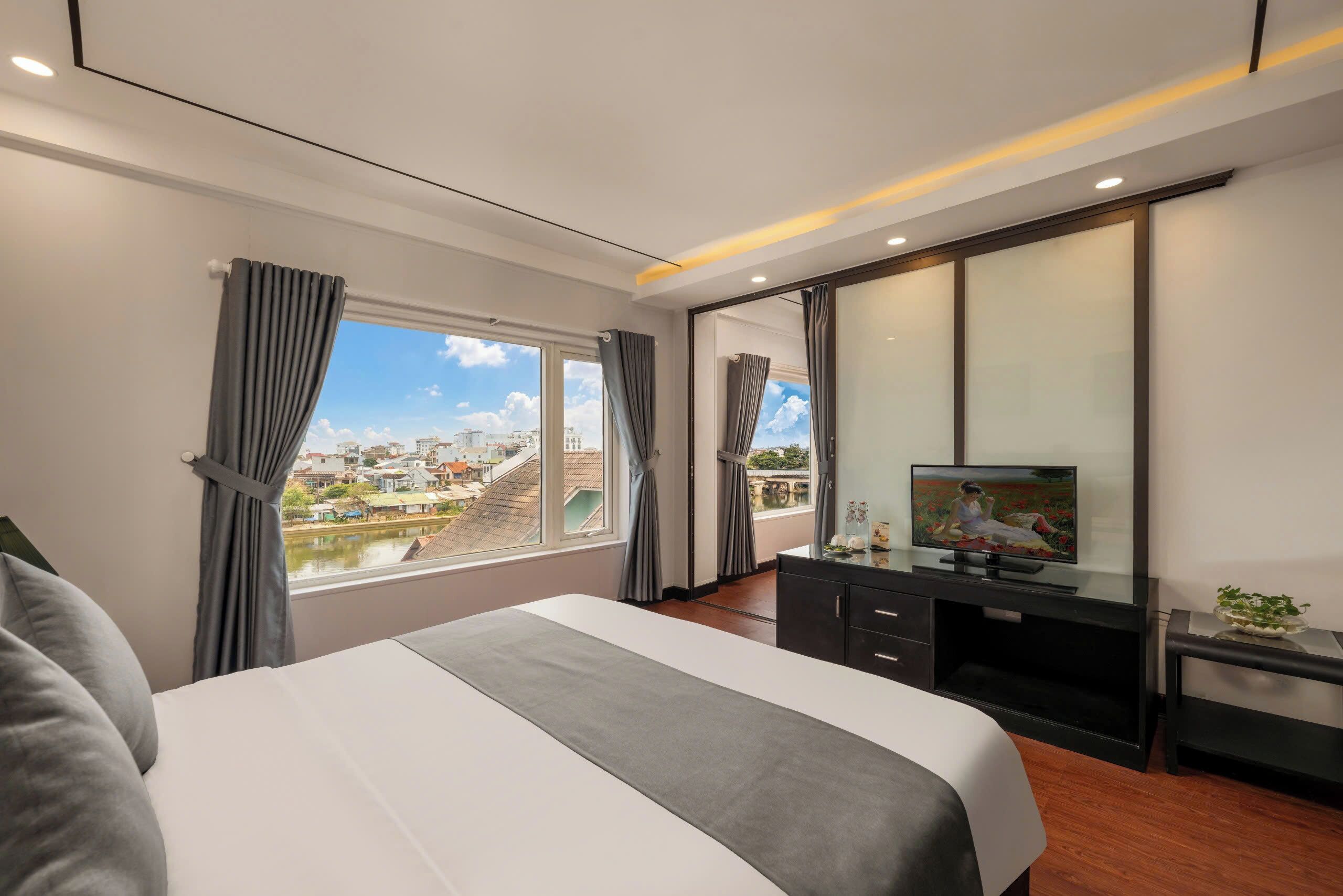 A modern hotel room with a large bed, a flat-screen TV, a desk, and a view of a cityscape through the window.