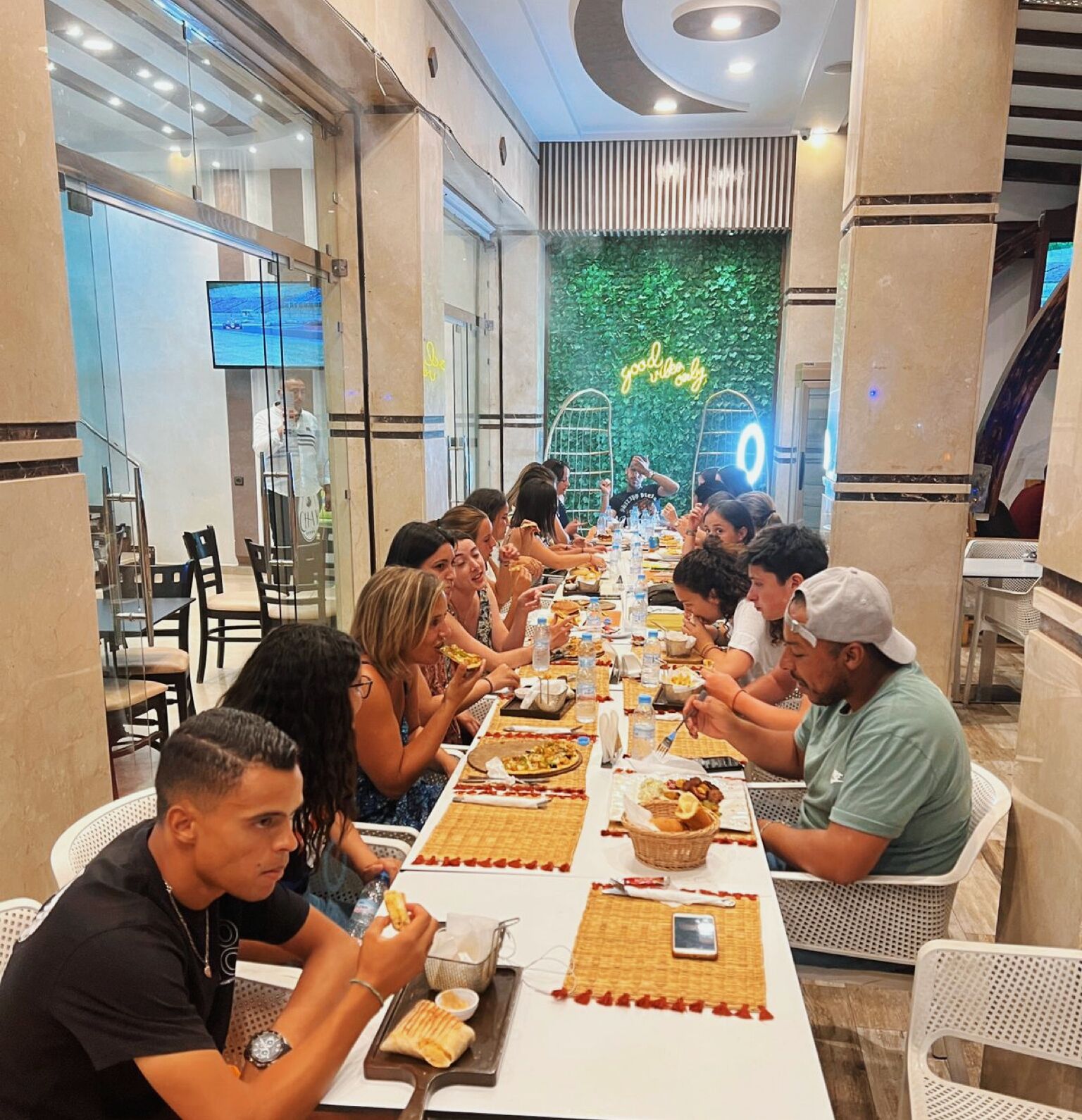 A group of people dining at a long table in a modern restaurant with a green leafy wall and a neon sign.