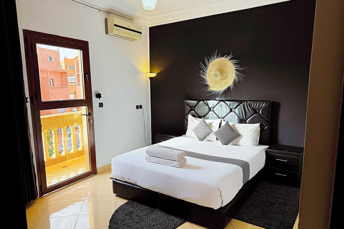 Habitación de hotel con cama, mesita de noche, lámpara y vistas al balcón exterior.