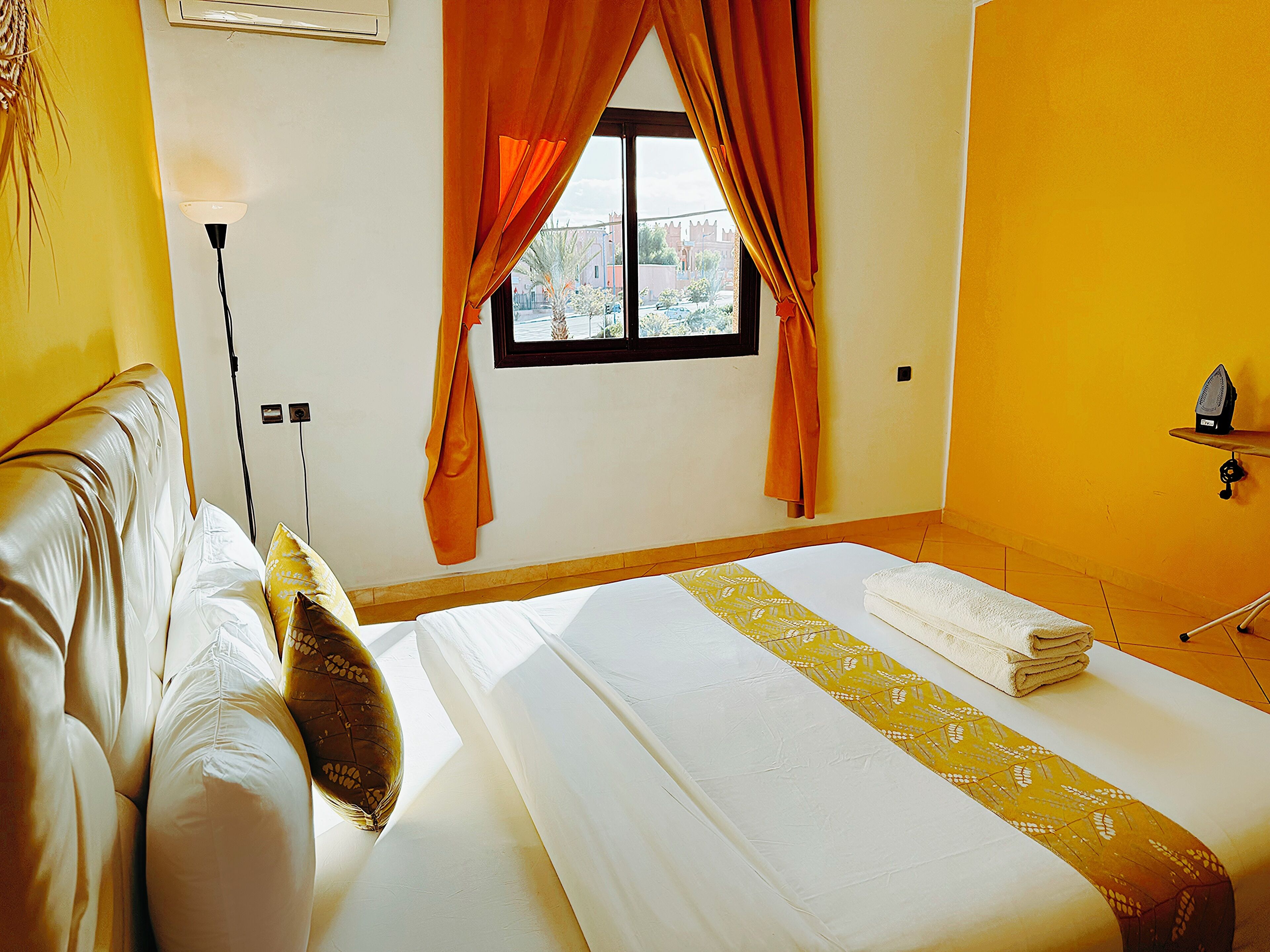 A hotel room with a bed, a window with curtains, a bedside table with an iron, and a wall-mounted air conditioning unit.