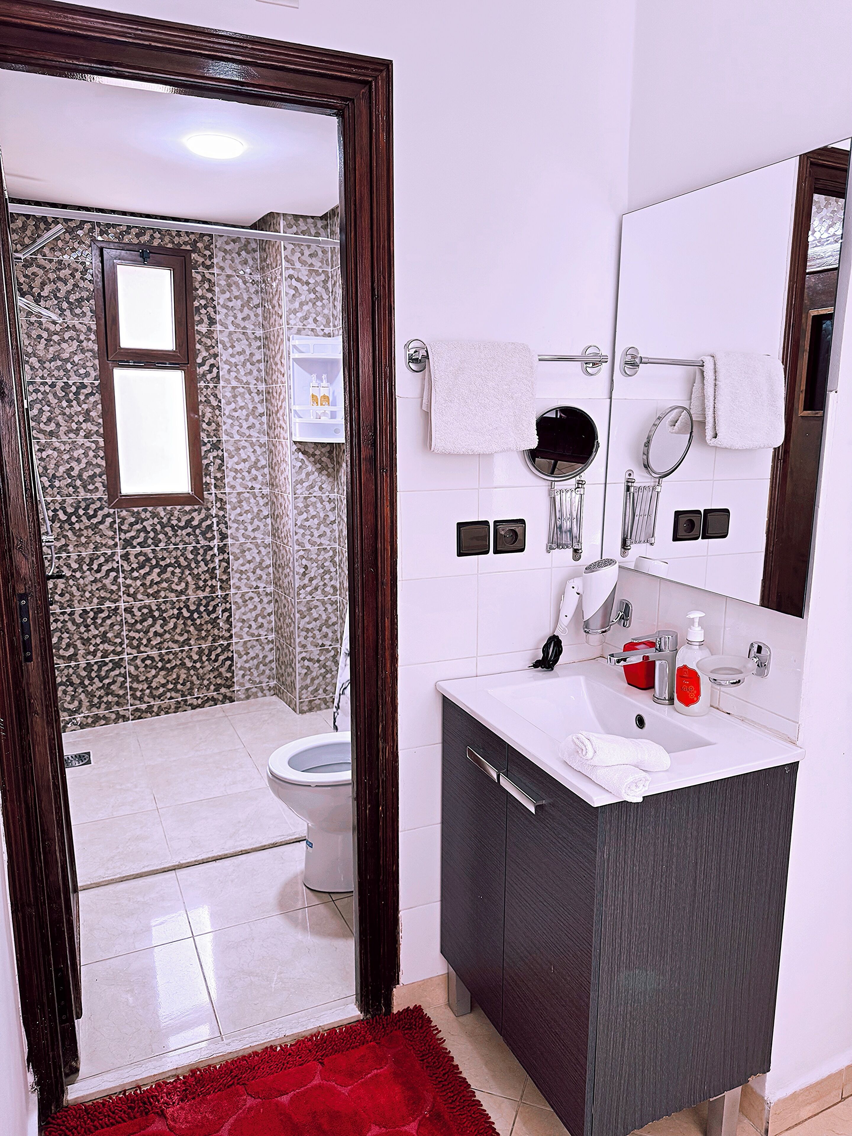 A bathroom with a toilet, sink, and mirror. There is a red rug on the floor and a shower visible in the background.