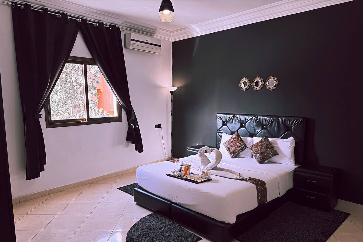 Habitación de hotel con cama, ventana con cortinas, mesita de noche y aire acondicionado instalado en la pared.