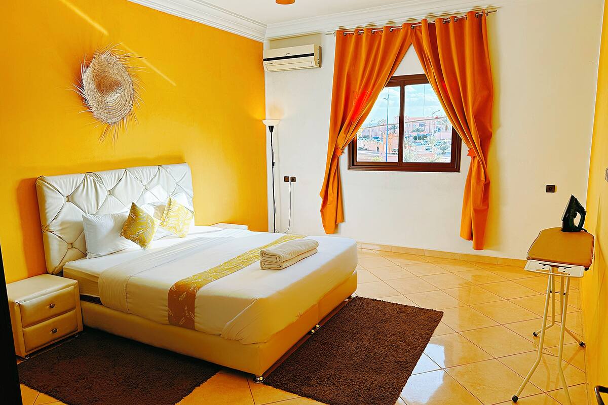 Habitación de hotel con cama, mesita de noche, tabla de planchar y ventana con cortinas.