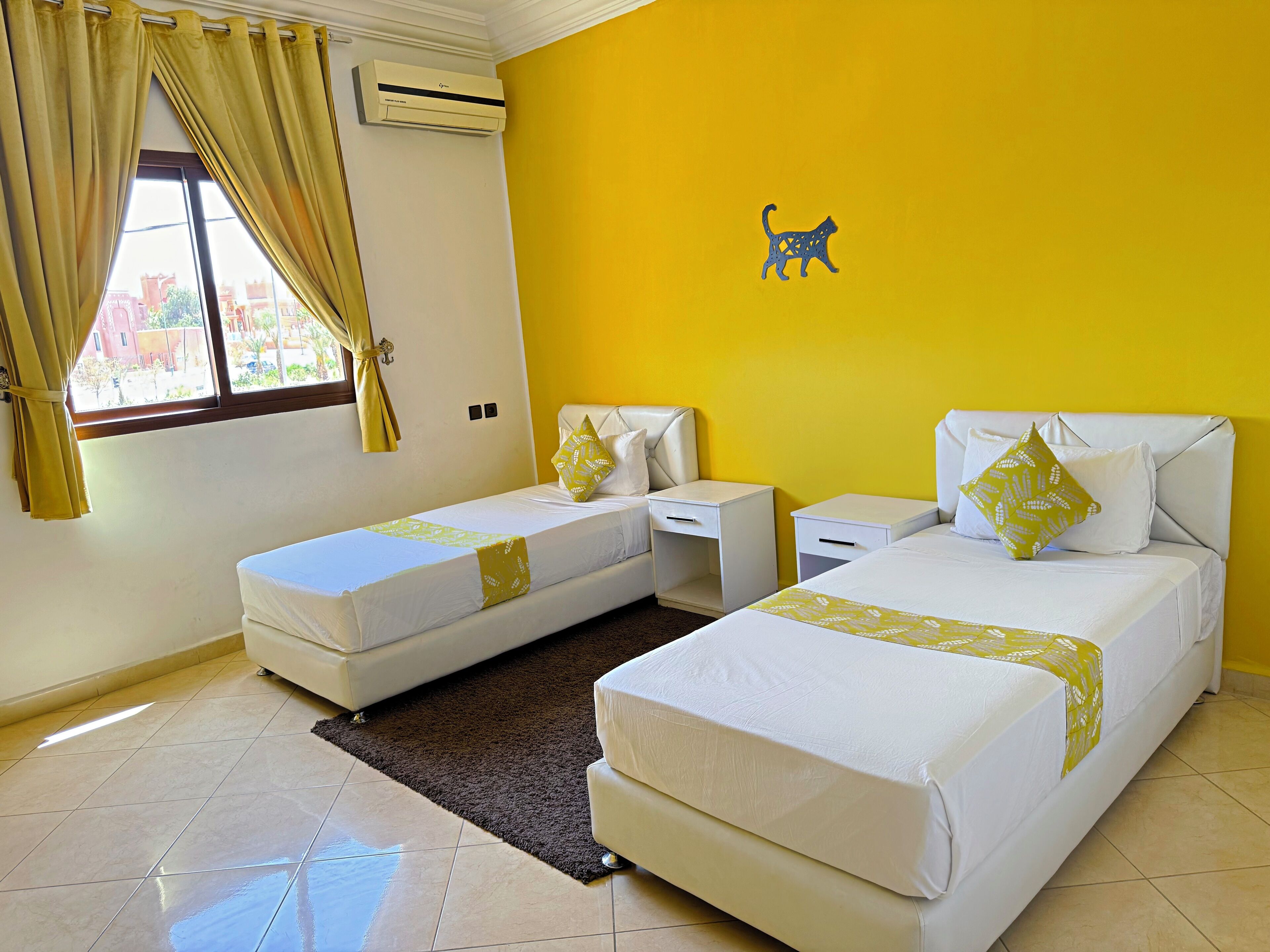 A hotel room with two beds, a window with curtains, an air conditioner, and a wall-mounted cat decoration.