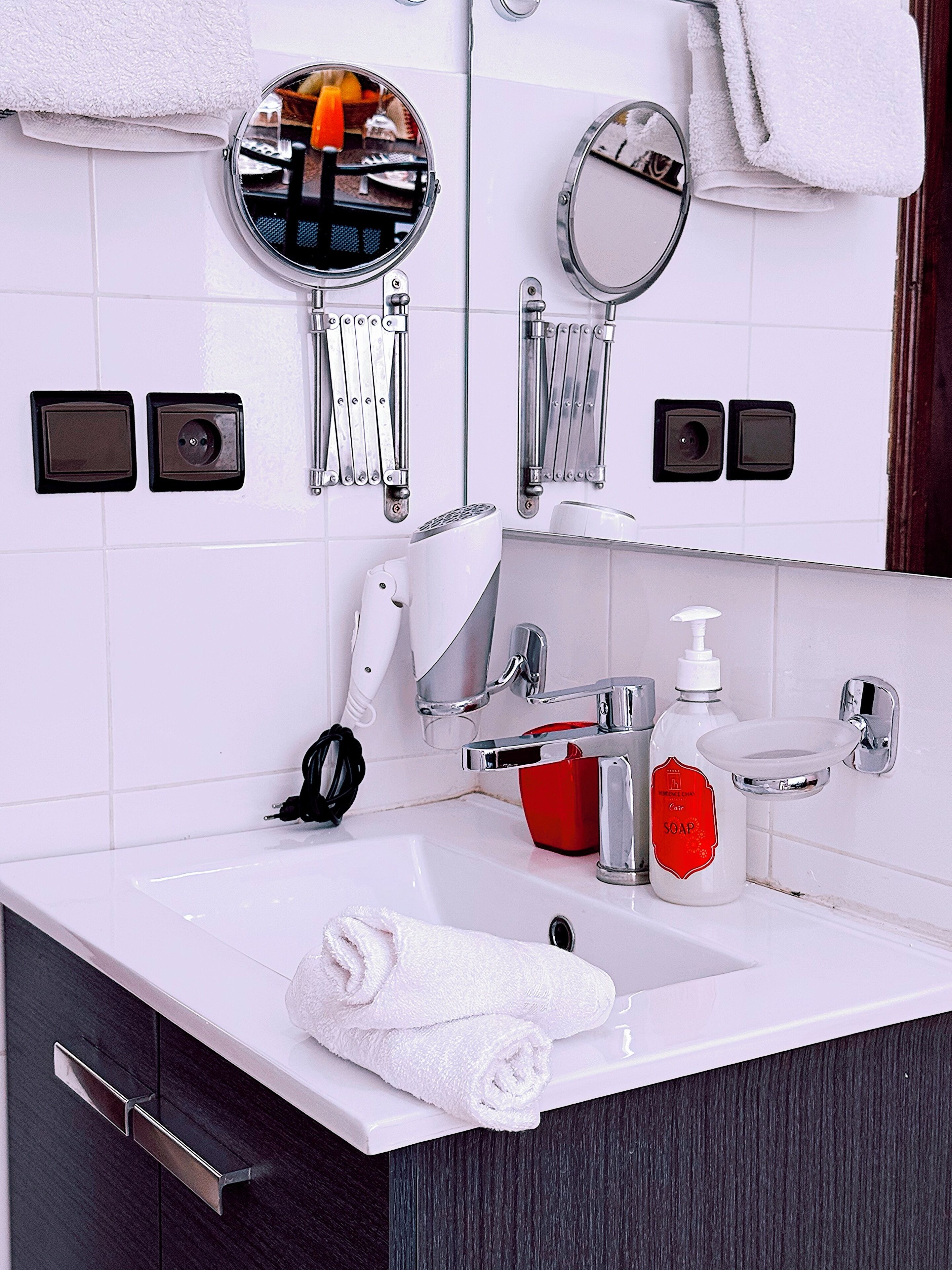 A modern bathroom with a white sink, chrome fixtures, a red soap dispenser, and two mirrors.