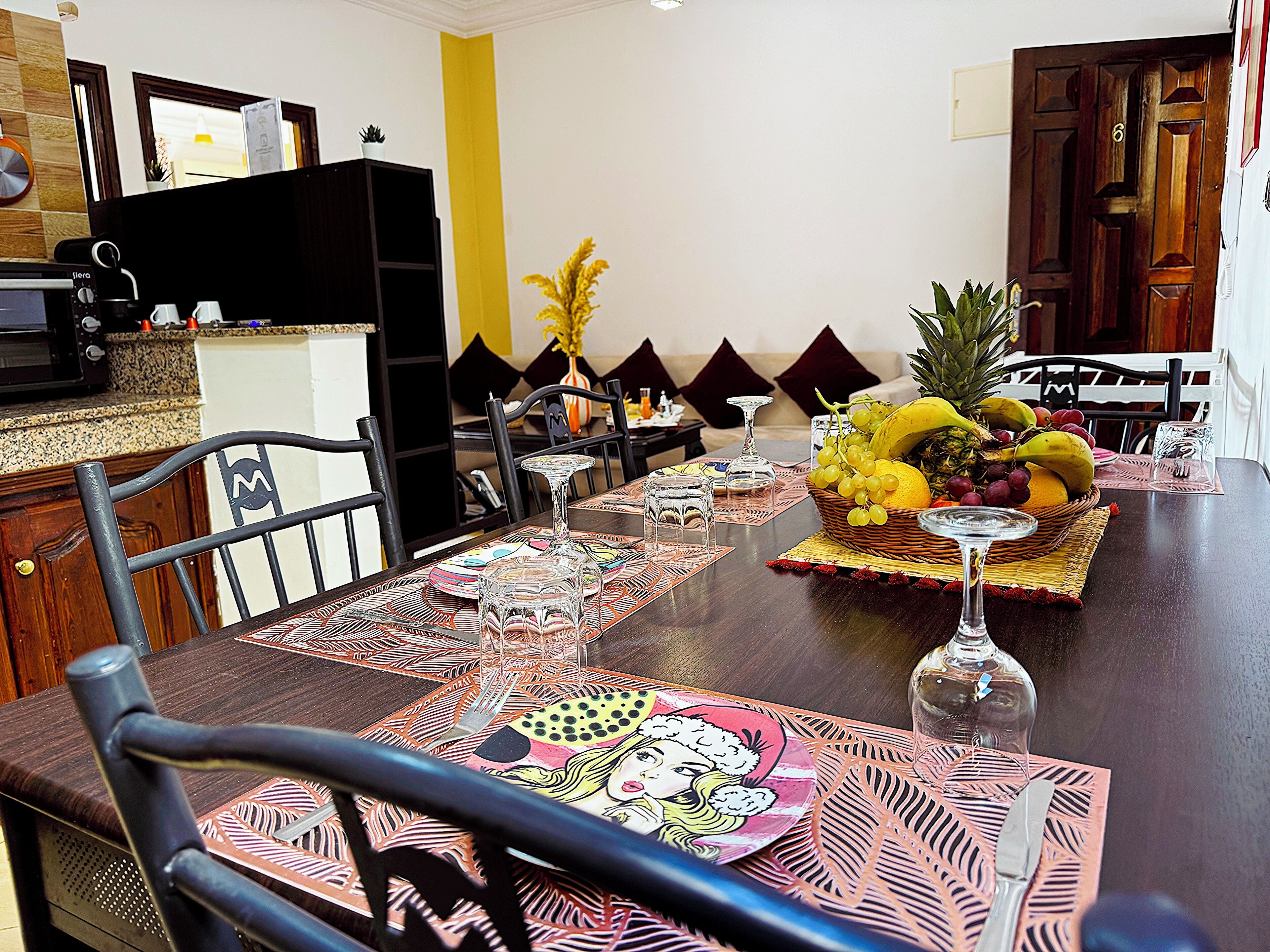 A dining area with a table set for four, featuring a fruit basket, wine glasses, and a decorative centerpiece.