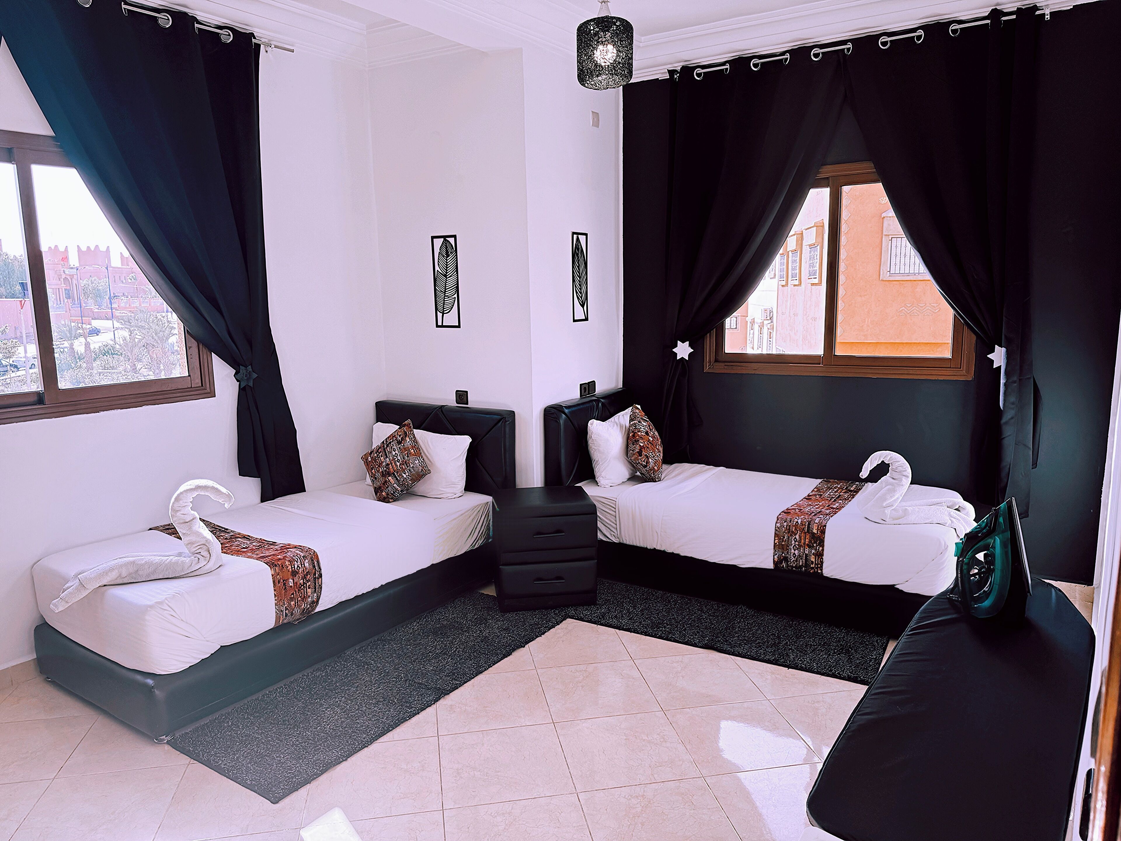 A hotel room with two beds, a small seating area, a window with curtains, and a view of buildings outside.