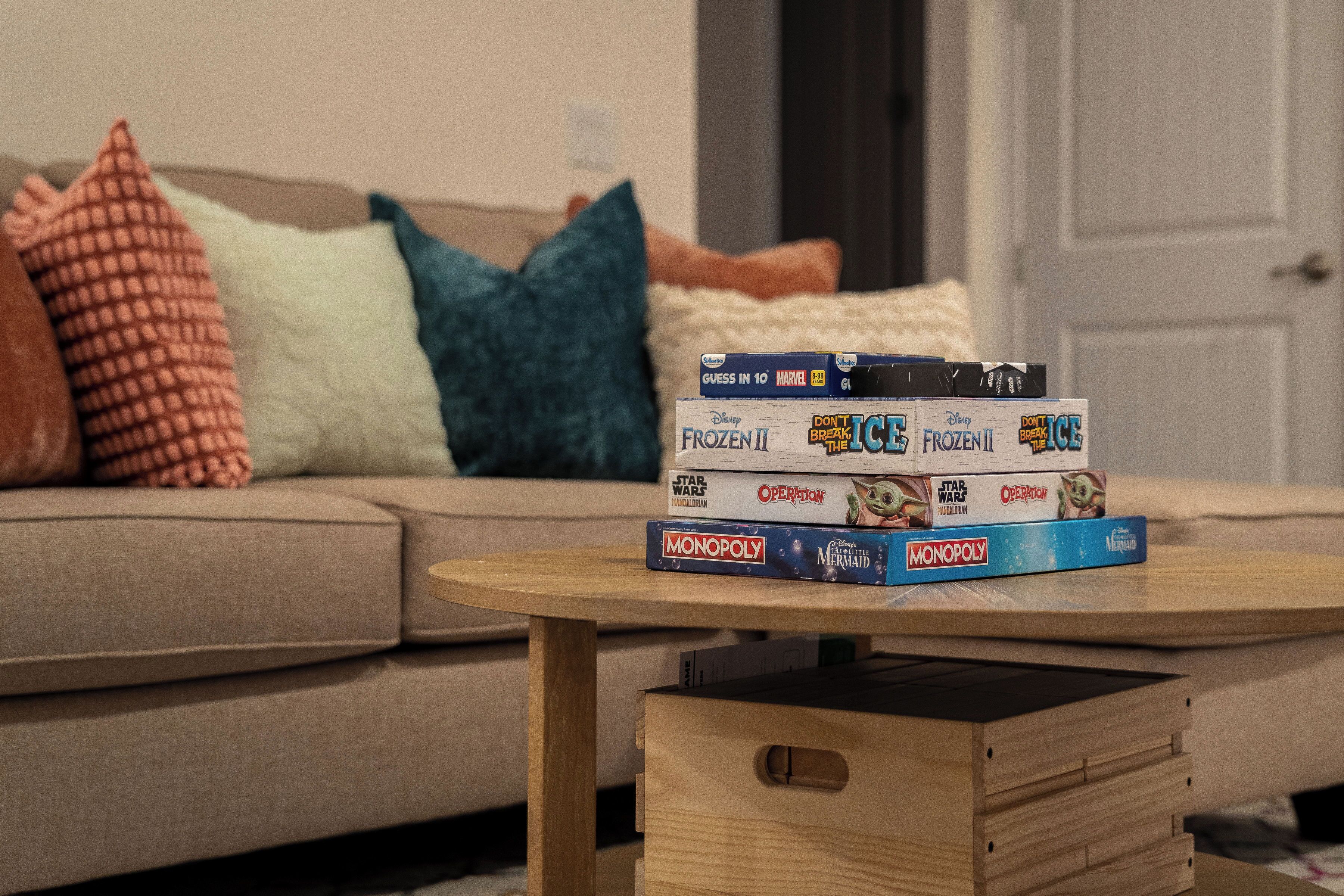 A stack of board games on a small wooden table in a living room with a sofa and pillows in the background.