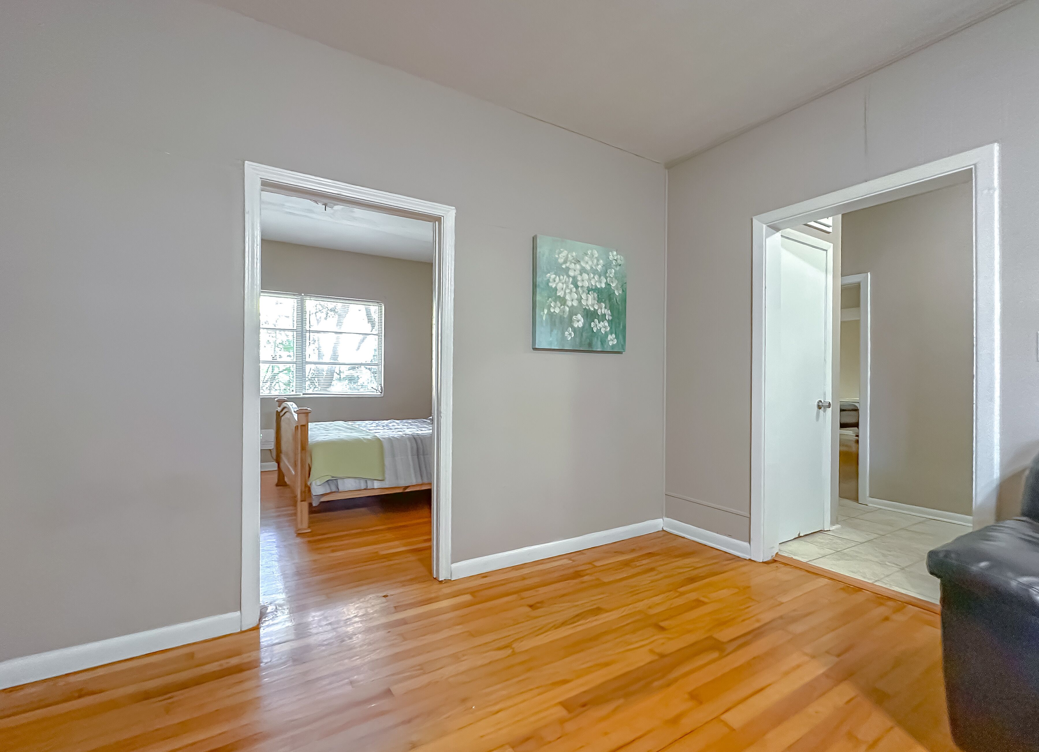 Une pièce avec un plancher en bois, un lit recouvert d’une couverture verte, une fenêtre avec des stores et un tableau accroché au mur.