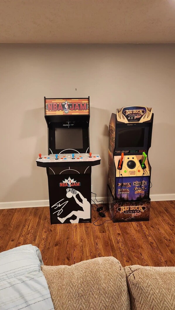 Two arcade machines, one labeled NBA JAM and the other Big Buck Hunter, placed against a wall with a wooden floor.