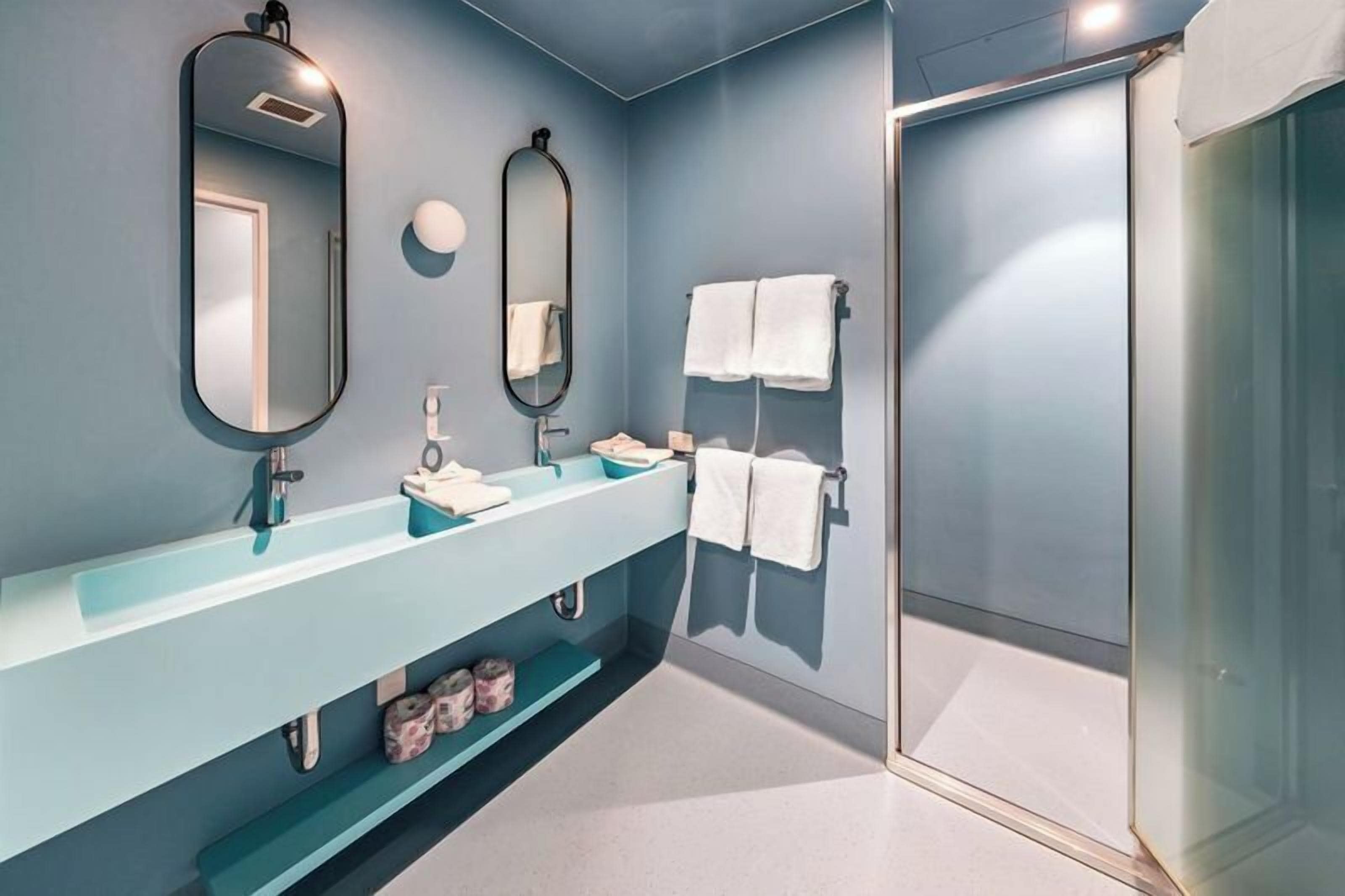Un baño moderno con un tocador azul claro, dos espejos, cabina de ducha de vidrio y toallas blancas.