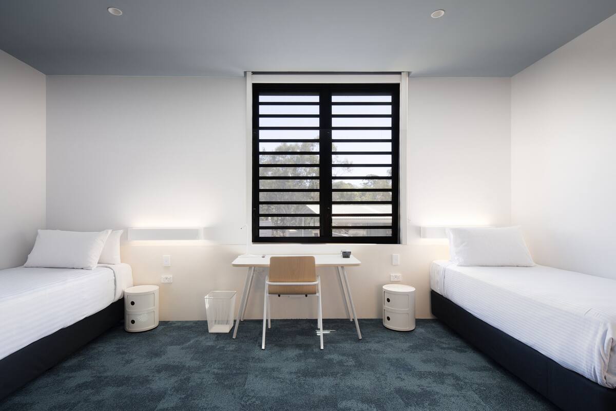 Une chambre d’hôtel avec deux lits, un bureau et une chaise. Il y a deux tables de chevet et une fenêtre avec des stores.