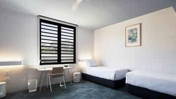 A modern hotel room with a desk, two beds, a window with blinds, a ceiling fan, and a framed picture on the wall.
