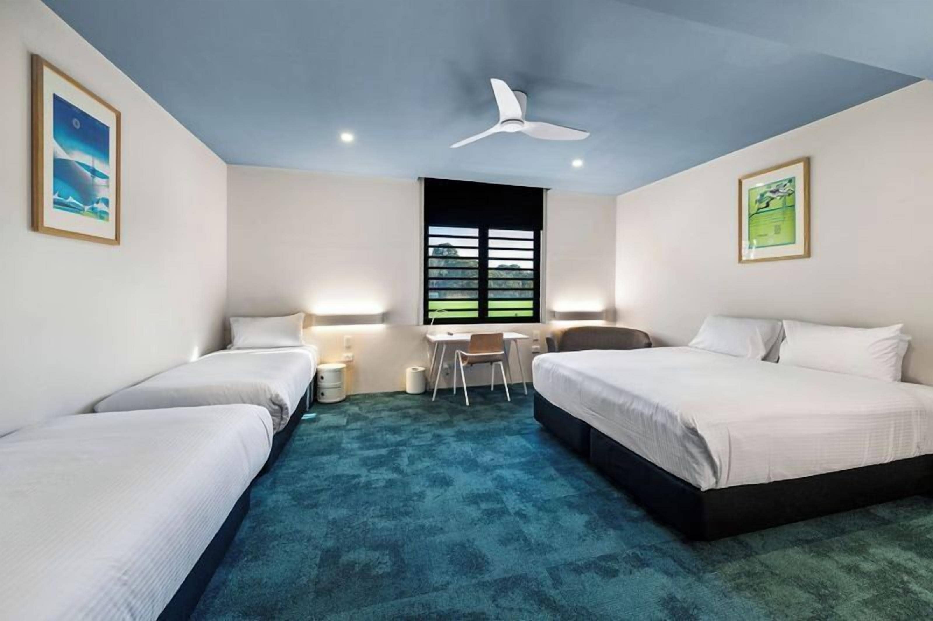 A hotel room with two beds, a desk, a chair, a ceiling fan, and a window with a view.