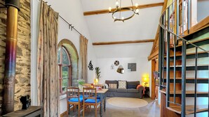Uma sala de estar acolhedora com uma parede de pedra, lareira, área de jantar com cadeiras de madeira, sofá e escada com guarda-corrimão de madeira.