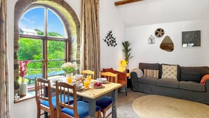 Uma área de jantar com uma mesa posta para dois, um sofá e uma grande janela com vista para o verde.