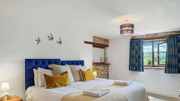 Un dormitorio con cabecera azul, cama con ropa de cama blanca y amarilla, mesita de noche con lámpara, cómoda, chimenea de piedra, lámpara en el techo y ventana con cortinas.