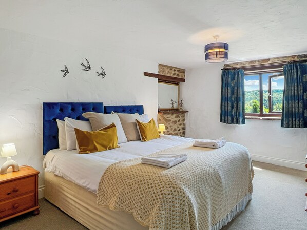 Quarto com cabeceira azul, cama com roupa de cama branca e amarela, mesa de cabeceira com abajur, cômoda, lareira de pedra, luminária no teto e janela com cortinas.