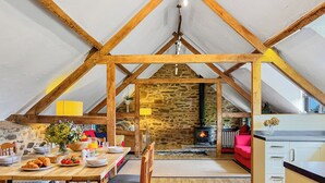 Acogedor dormitorio en ático con pared de piedra, vigas de madera, chimenea, mesa de comedor con sillas y zona de cocina.