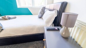 Una cama bien hecha con un control remoto en una mesita de noche, una lámpara y una toalla enrollada en el suelo.