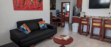 A living room with a black sofa, a round coffee table, and a ceiling fan. There is a dining area with wooden chairs and a TV mounted on the wall.