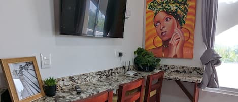Cozinha com televisão na parede, um quadro sobre o balcão e uma janela com cortinas.
