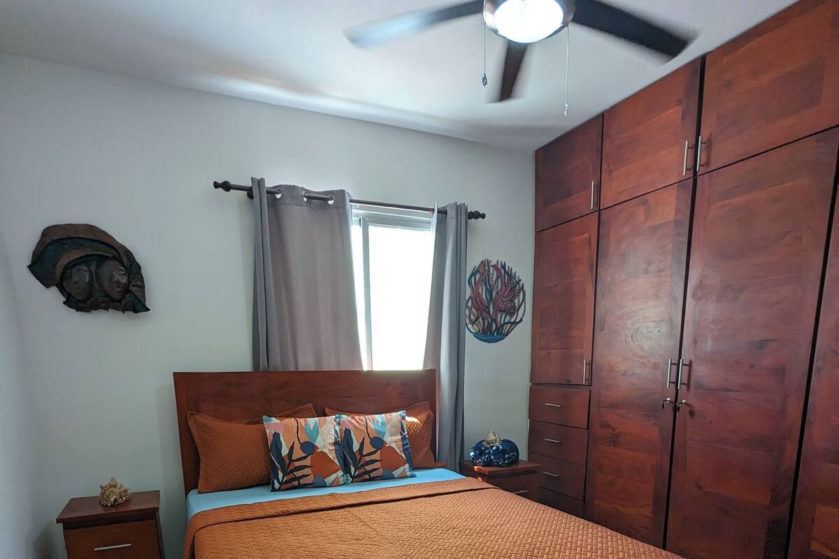 Un dormitorio con cama, mesita de noche, armario, ventilador de techo y ventana con cortinas.