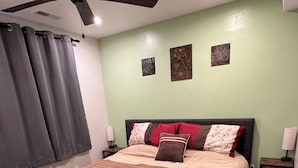 A bedroom with a bed, a ceiling fan, a nightstand with a lamp, and a wall with two framed pictures.