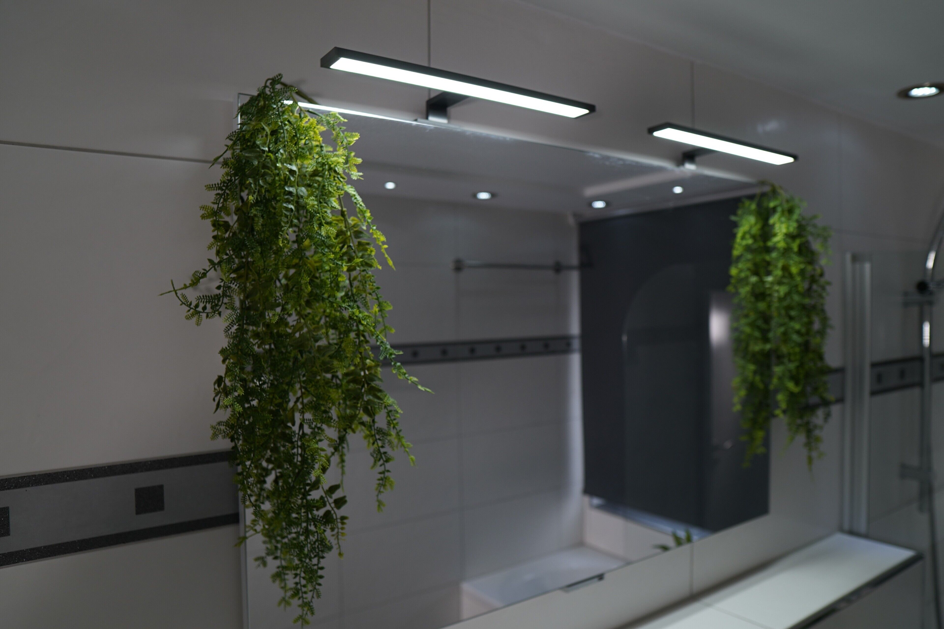 A modern bathroom with a large mirror, hanging plants, and recessed lighting.