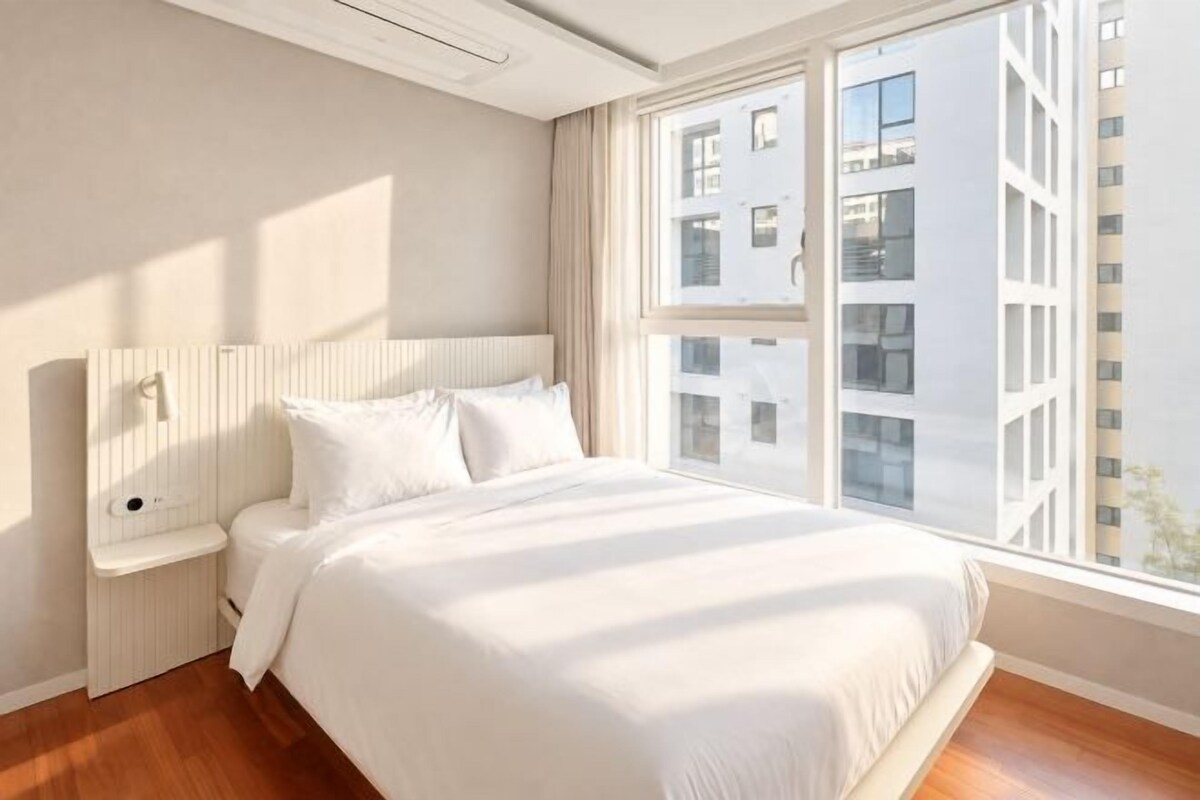 A hotel room with a large bed, a bedside table, a window with a view of buildings, and a ceiling light.