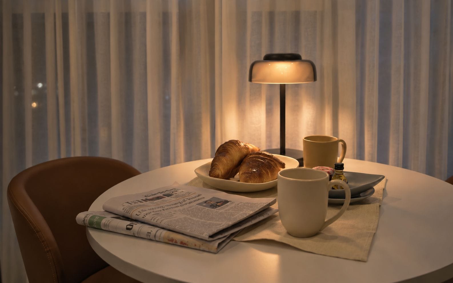 Una mesa redonda con un periódico, un plato de cruasanes, una taza y una lámpara.